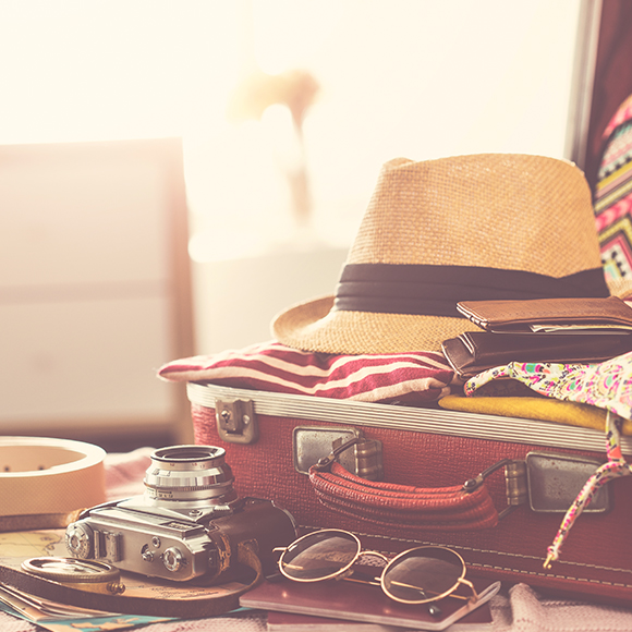travel-2-product Packed suitcase laying open on bed, complete with a straw hat, camera and sunglasses to promote travel insurance by Evalee Insurance Brokers